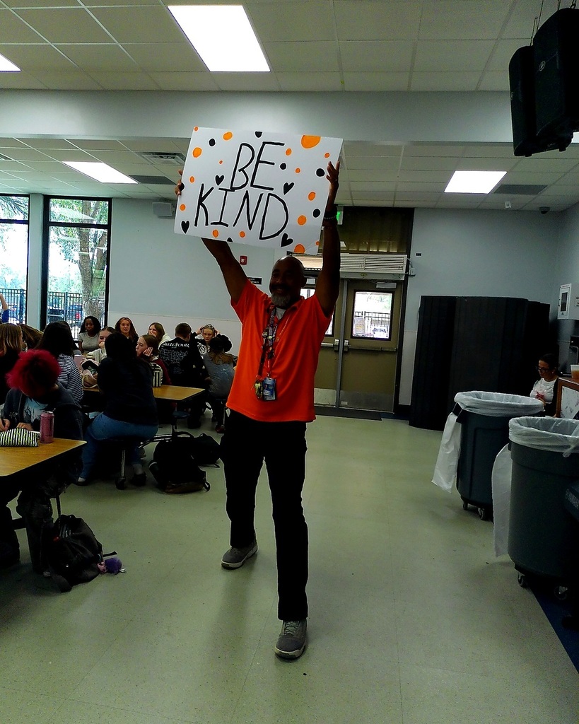 Man holding up a sign reading "BE KIND" over his head in a room with tables.