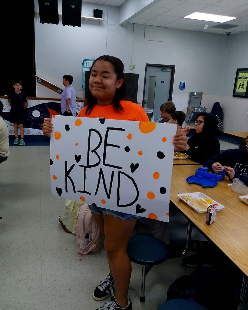 Student holds up white sign with orange dots and black text, "Be Kind," in a classroom with tables.