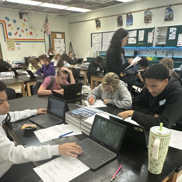 Students in a classroom, seated at tables, using laptops and writing in notebooks, with a woman standing at a nearby desk.
