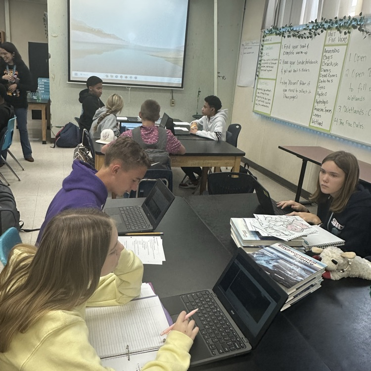 Several students in a classroom sit at tables using laptops and reading books, one student writing.