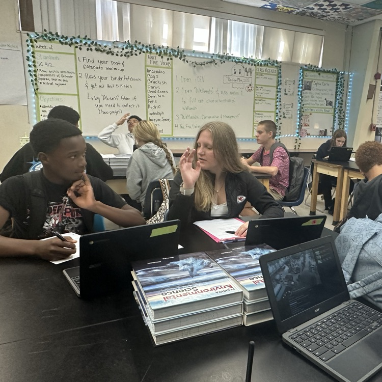 Students seated in rows using laptops at tables, with a whiteboard and bulletin board in the background.