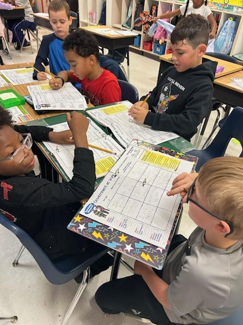 Students in a classroom, seated in blue chairs, are focused on a workbook and worksheets.