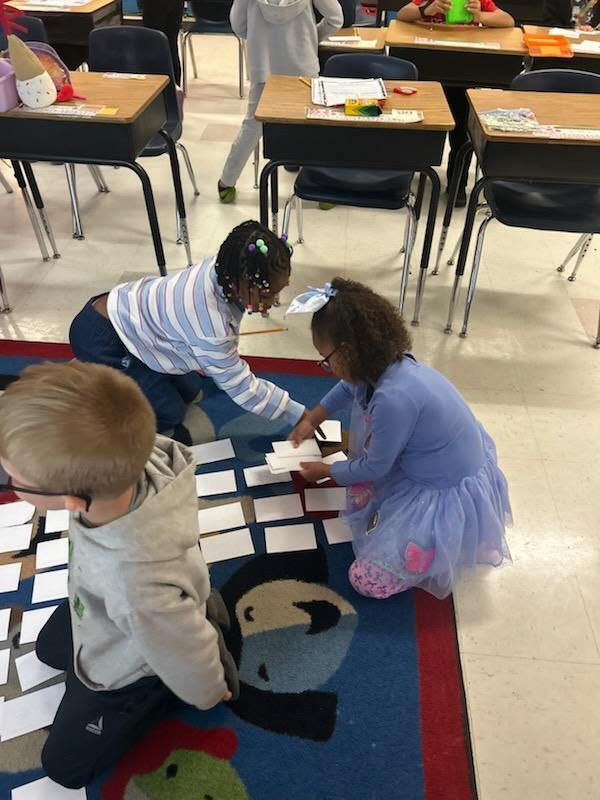 Three children in a classroom place paper rectangles on a rug with cartoon characters.