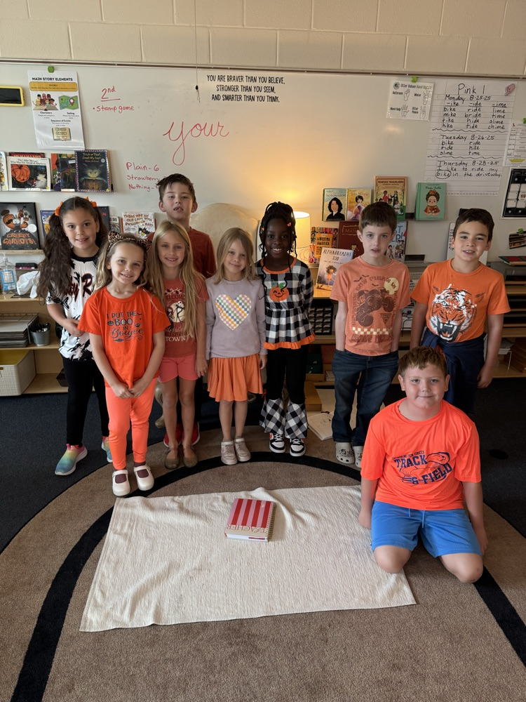 Students in a classroom stand and crouch around a book on a white cloth on a rug.