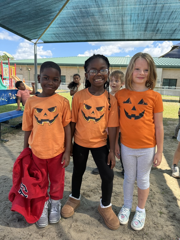 Four children in orange T-shirts with jack-o'-lanterns on them stand on a sandy surface.