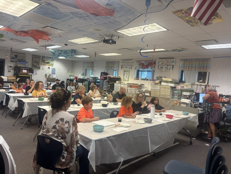 Classroom with tables covered in white cloth. People seated at tables. Woman standing in front of tables.