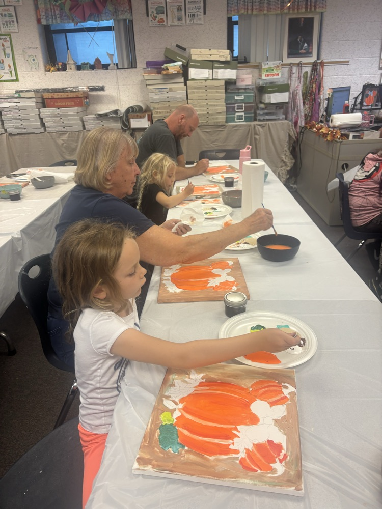 Three people, including two children, at a long table painting pumpkins with orange paint.