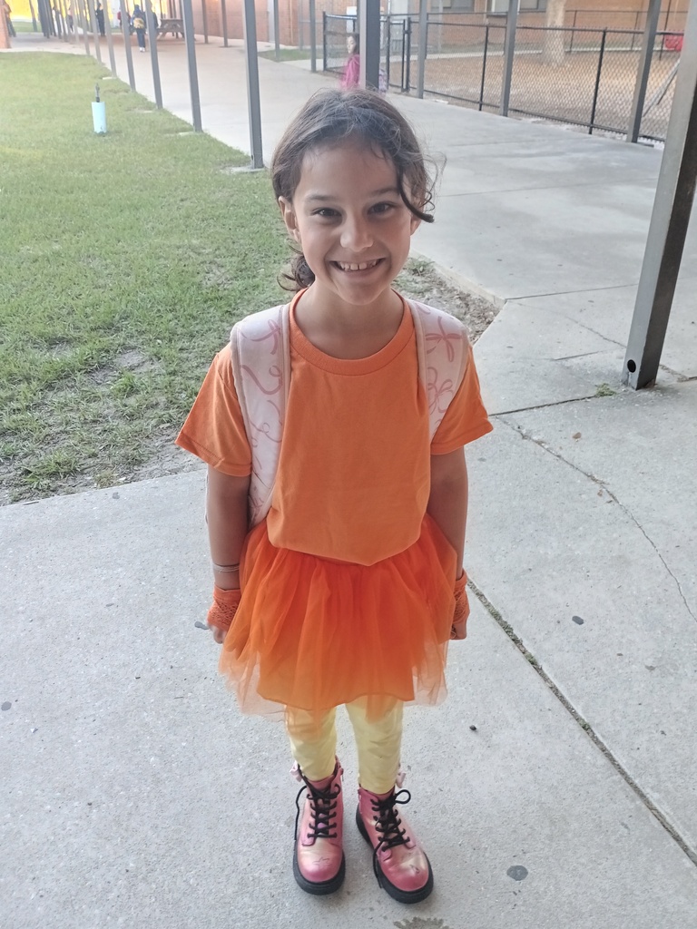 A child in an orange shirt, tutu, and pink boots stands on concrete with a smile.