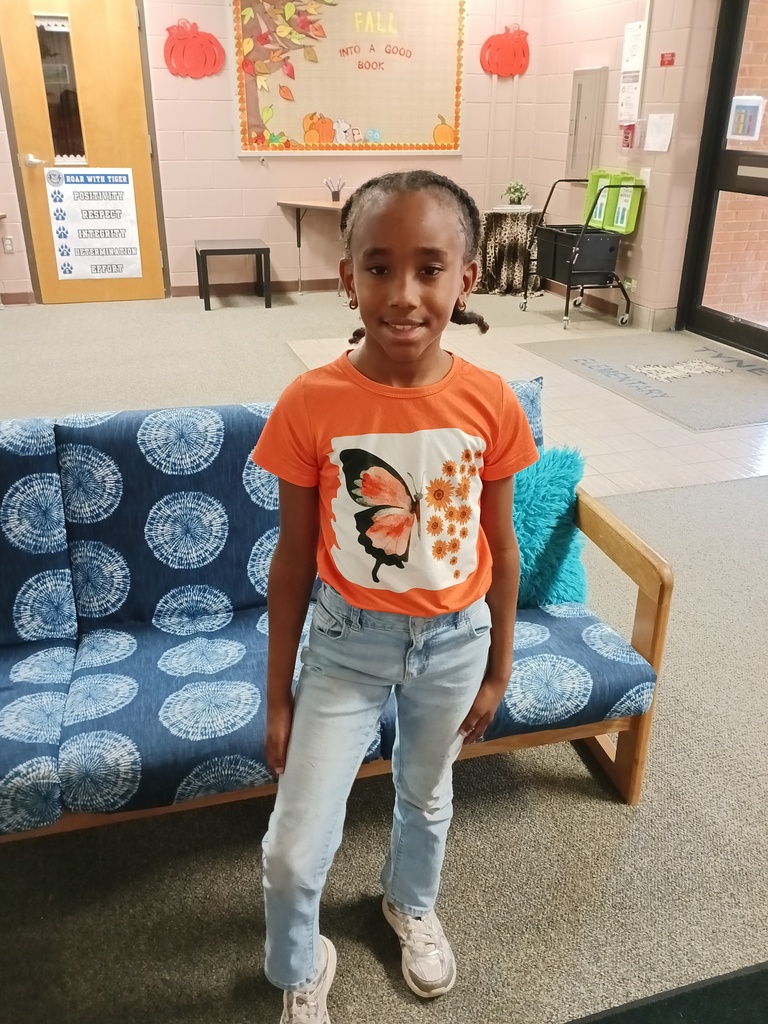 A young girl wearing an orange T-shirt with a butterfly design and white sneakers stands in front of a blue couch with circular patterns.