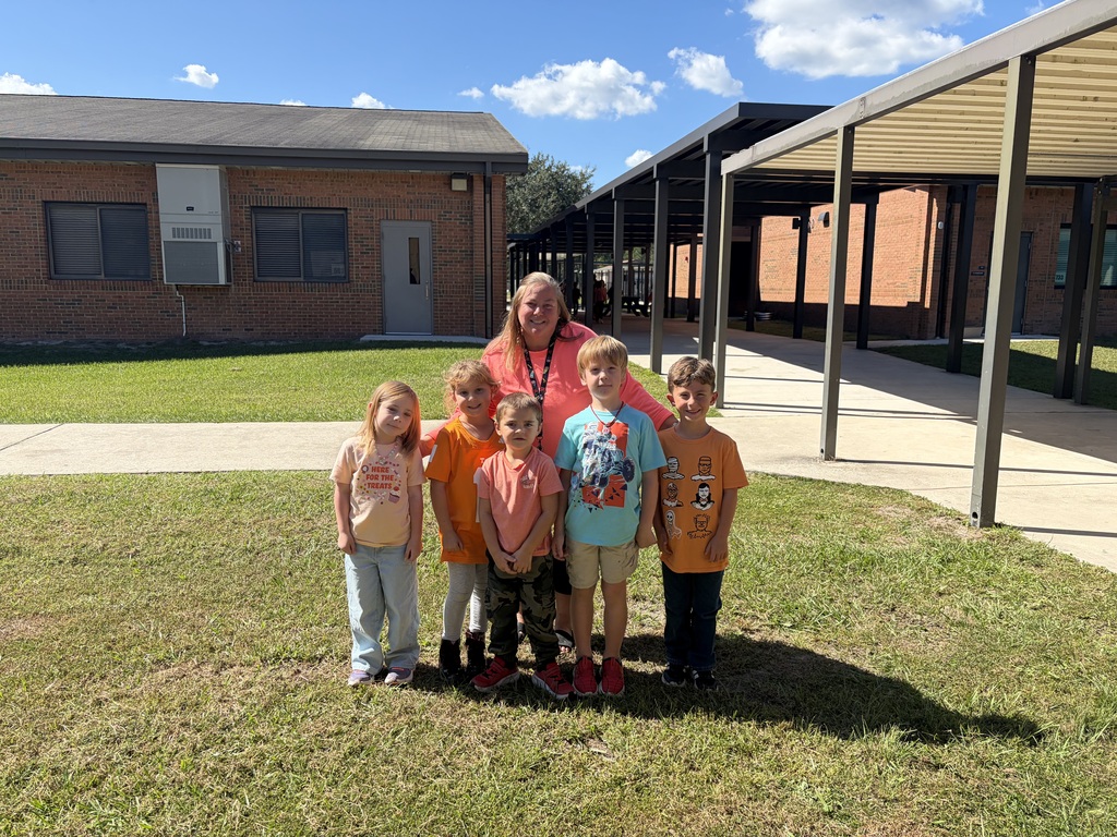 A group of children stands in front of a brick building with an adult, under a cloudy sky.