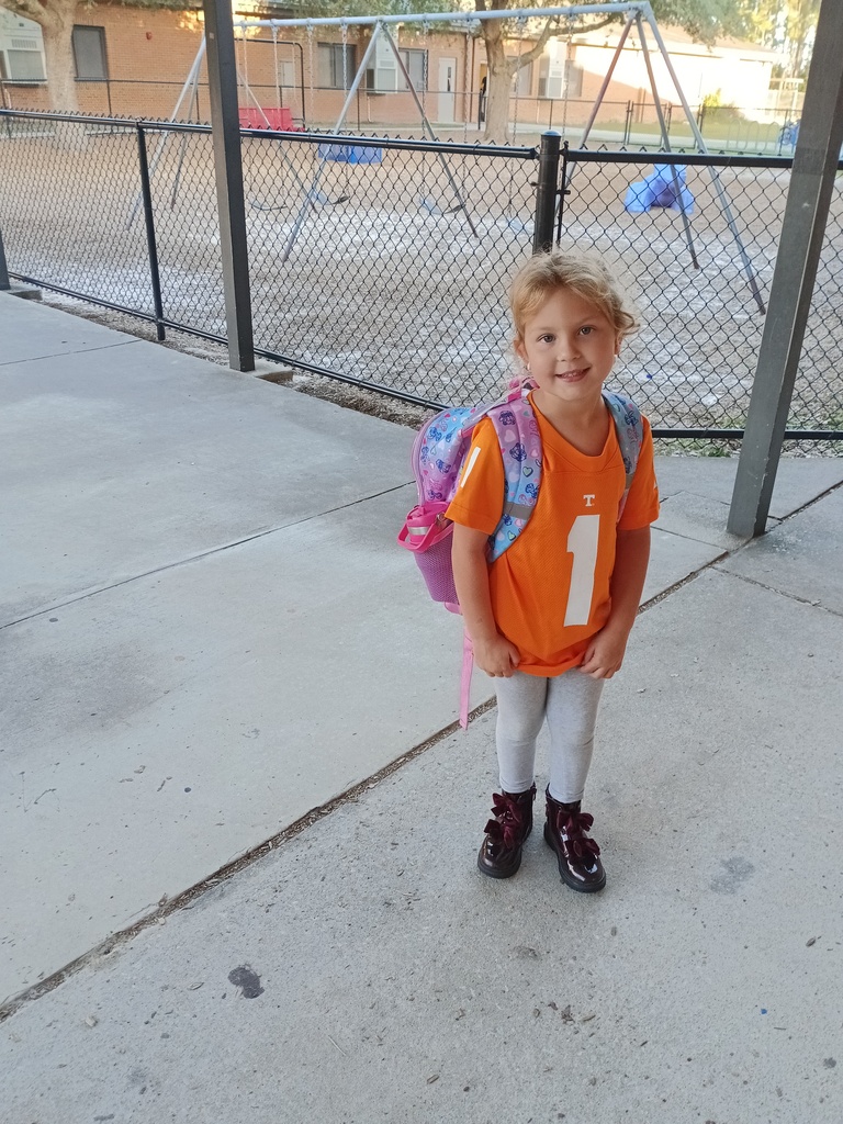 Child with backpack stands outside by fence; blurry playground and building in background.