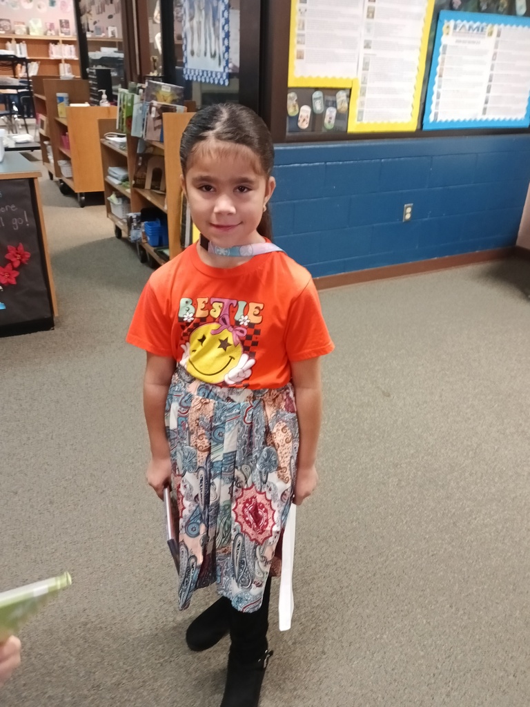 A young girl wearing an orange shirt stands in a classroom with bookshelves and bulletin boards.