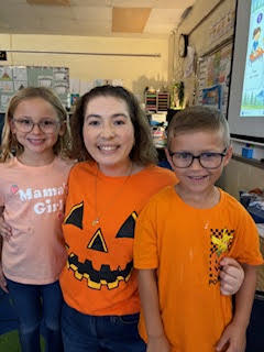 Three kids stand in a classroom, one girl in a pumpkin shirt, another in pink, and a boy in orange, all smiling.
