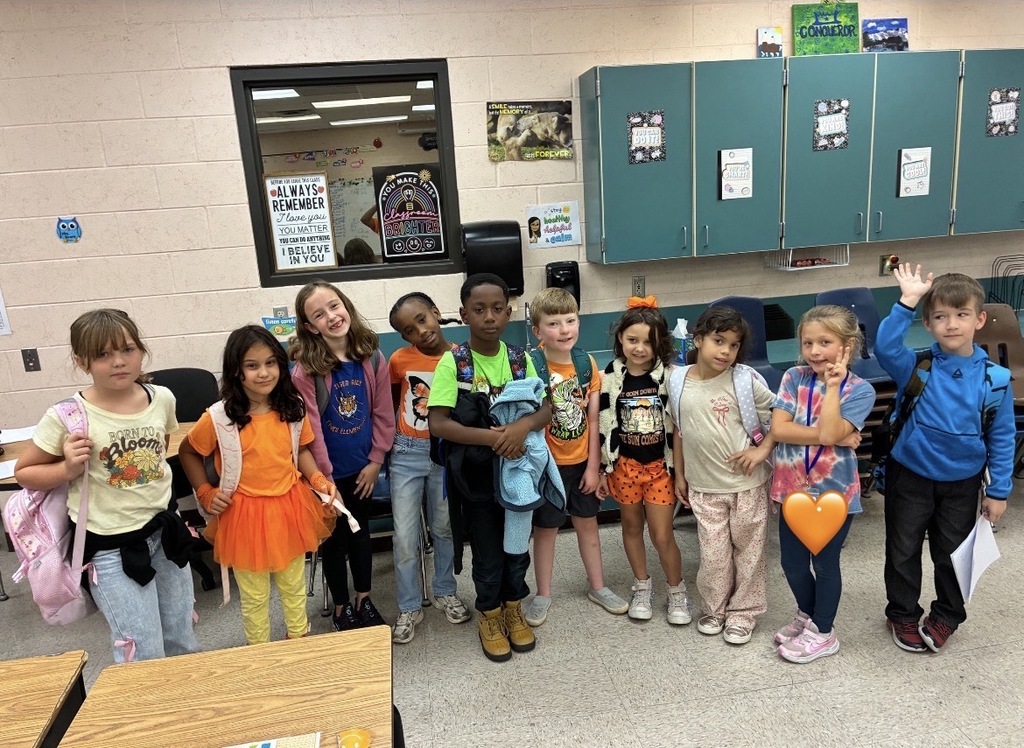 A group of young children stands in a classroom. Some wear backpacks. They face the camera.