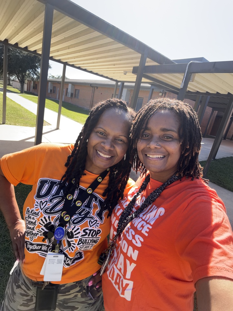 Two women standing outside a covered walkway, both wearing orange shirts and smiling at the camera.