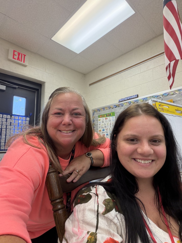 Two smiling women in a classroom with an American flag on a wall, one standing, the other seated.