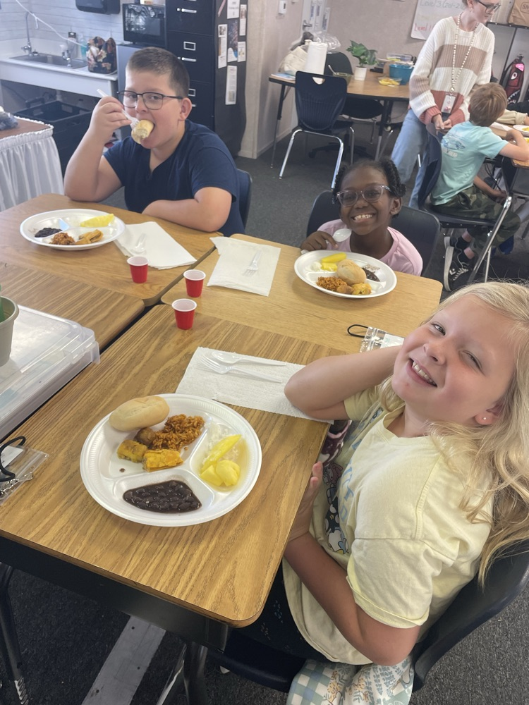 Several students seated at tables, eating. A girl smiling, holding her face. A boy eating with his mouth full.