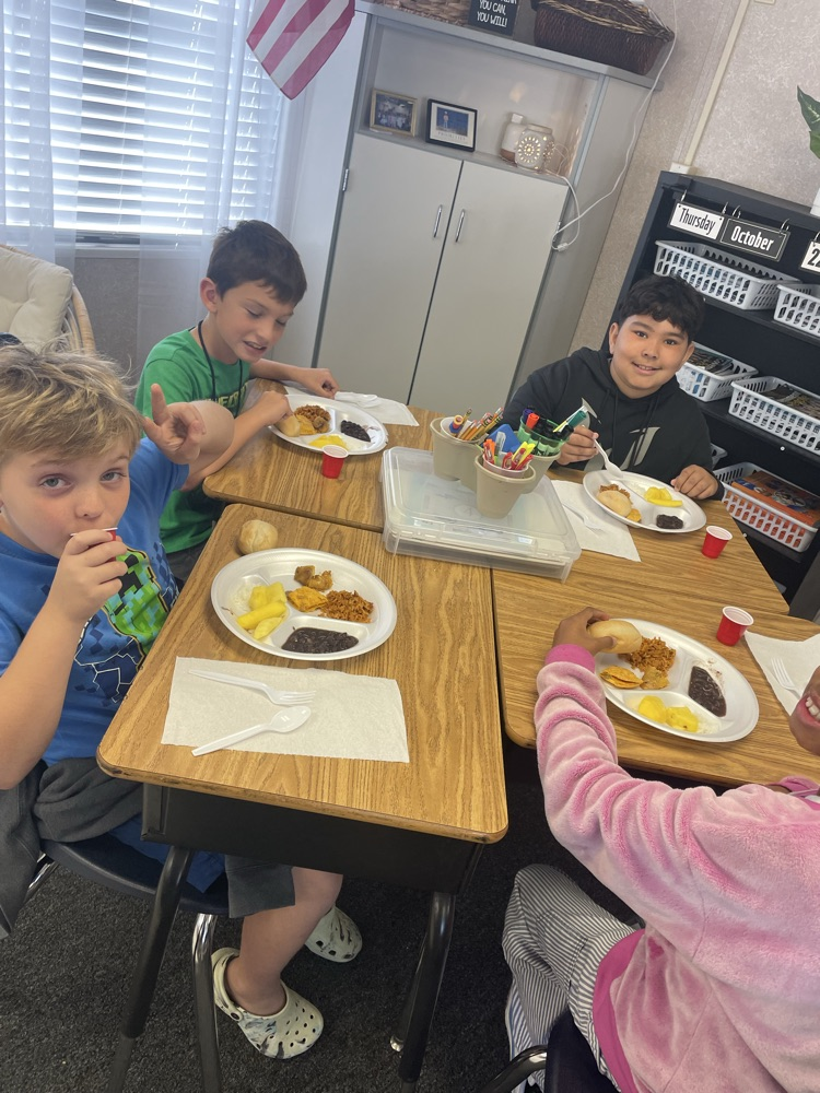Four students seated at tables in a classroom, eating food from paper plates and drinking from cups.