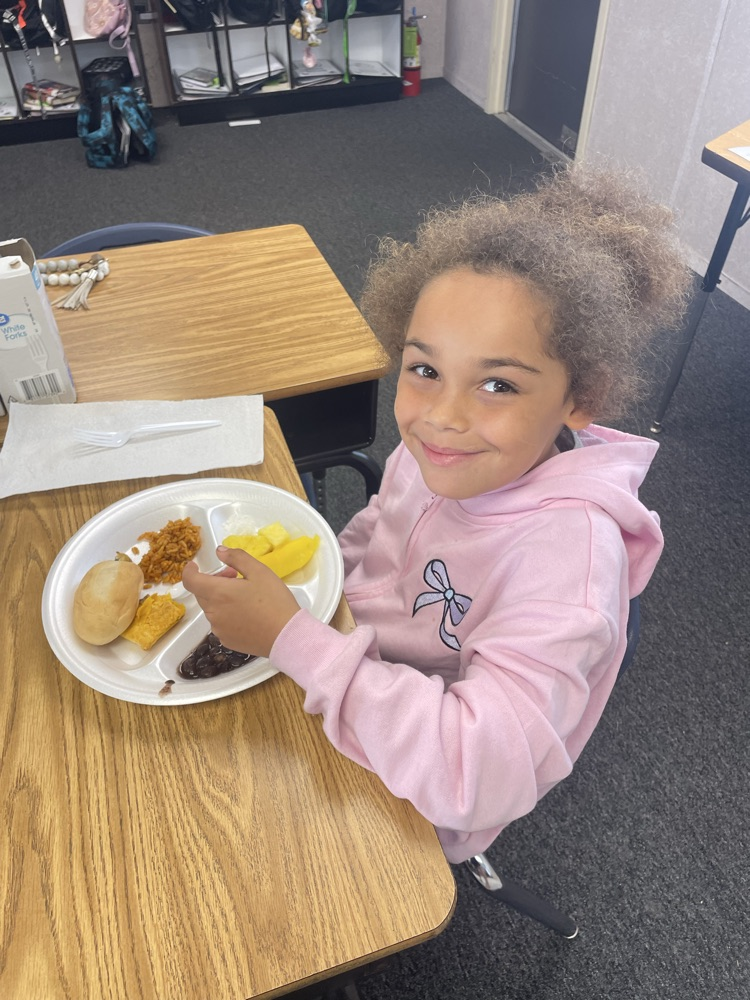 A child sits at a table with a plate of food, smiling. Behind them, a desk and chair.