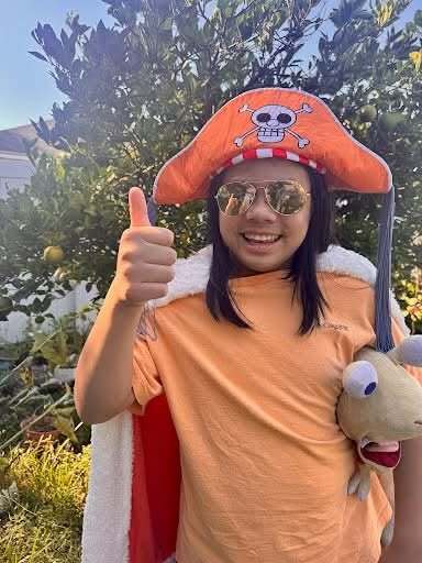 A person stands with a thumb up, wearing a skullcap and an orange shirt, holding a stuffed animal.
