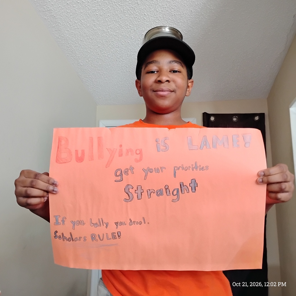 A boy holding a large orange poster with bold black writing. He is wearing a hat and an orange shirt.