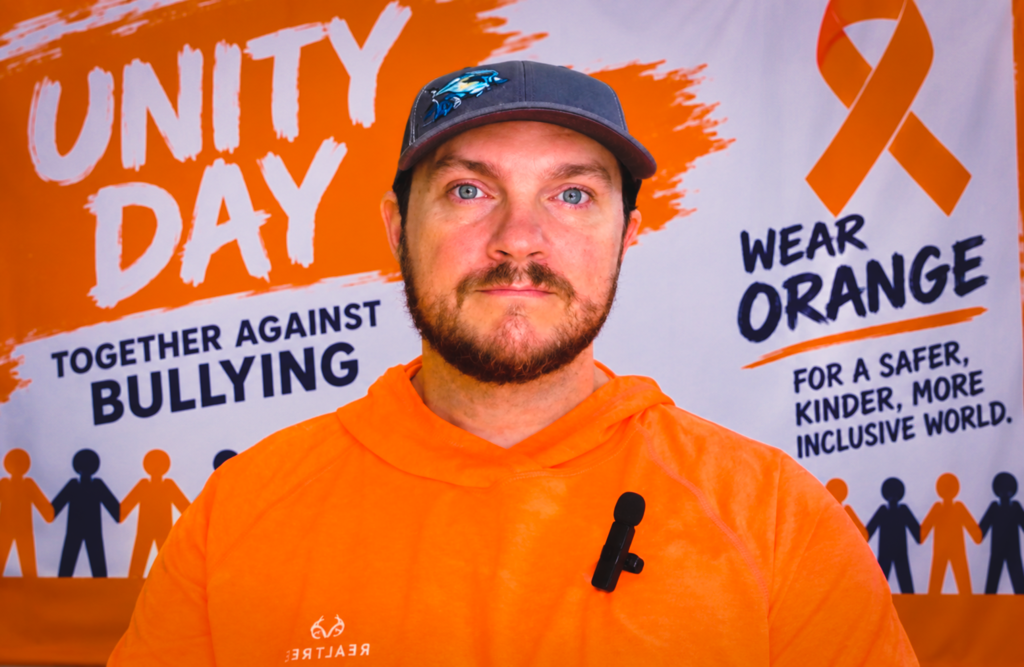 Man in orange shirt and hat standing in front of banner with silhouettes of people.