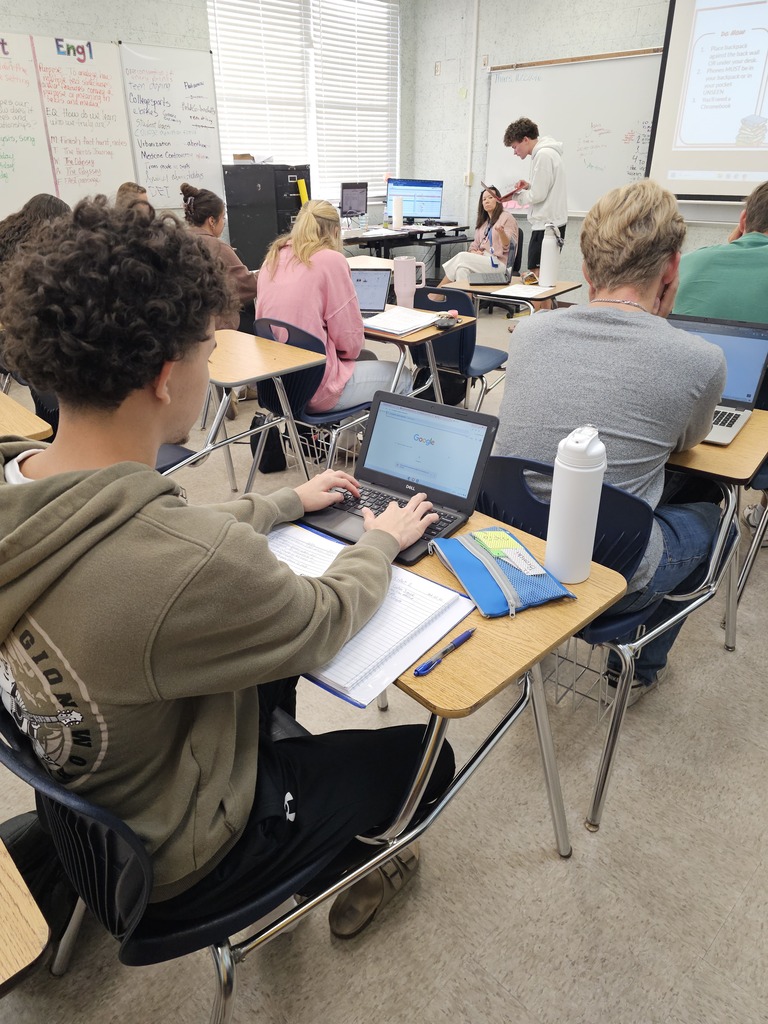 Classroom with students seated at desks, working on tablets, papers, and laptops. Light beige walls.