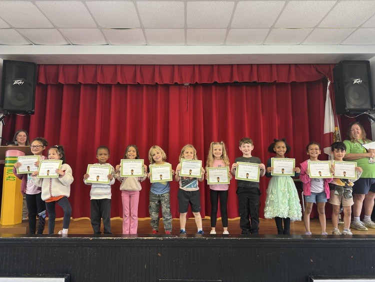 Children holding certificates stand on a stage with a red curtain backdrop, spotlights, and speakers.