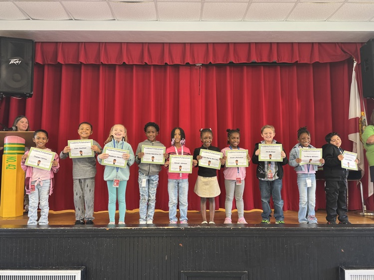 Nine children holding certificates standing in a row on a stage with a red curtain backdrop.