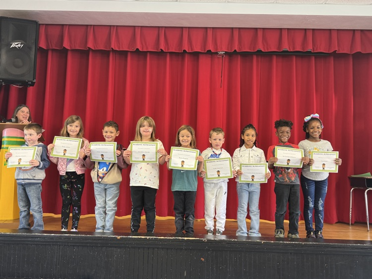Kids holding certificates stand on stage in front of a red curtain with a person in the background.