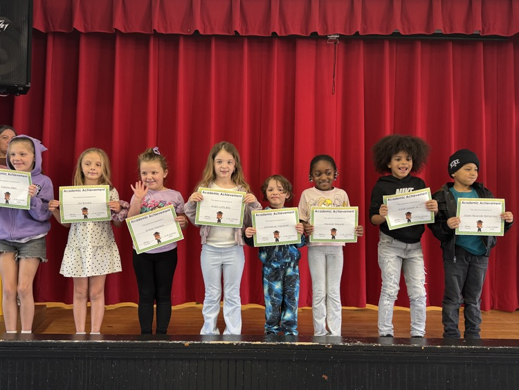A group of young students stands on stage, holding certificates, against a red backdrop.