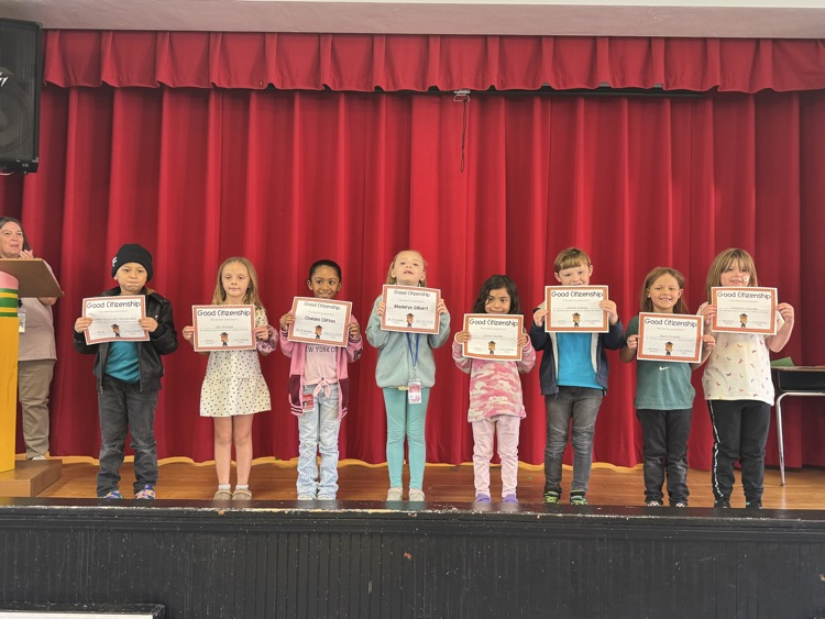 Eleven children stand on stage holding certificates, a person at the back with a book.