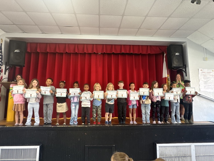 Children in a school auditorium holding certificates; a flagpole is to the left of the stage.