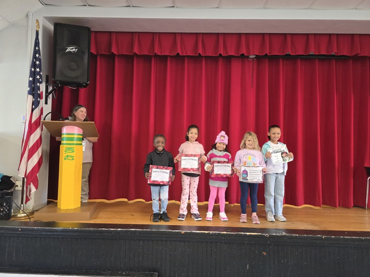 Children holding certificates stand on a stage with a red curtain and an American flag.
