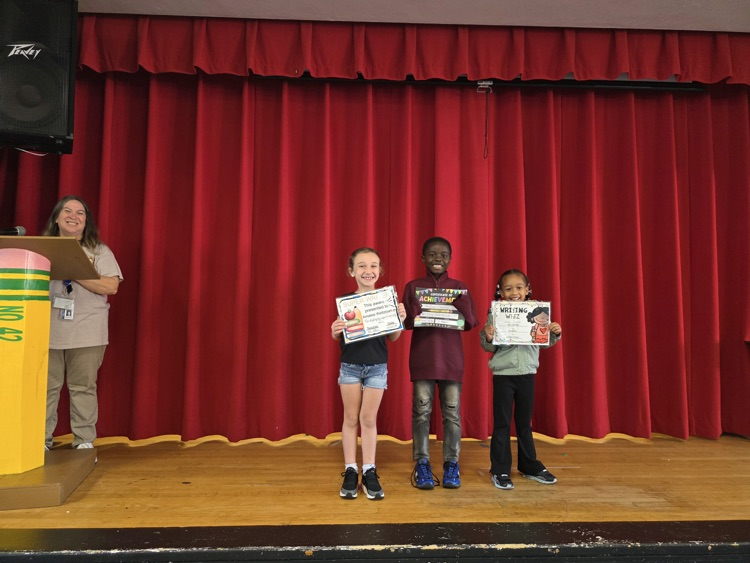 Three children with certificates stand on a stage; a woman in the background holds a microphone.