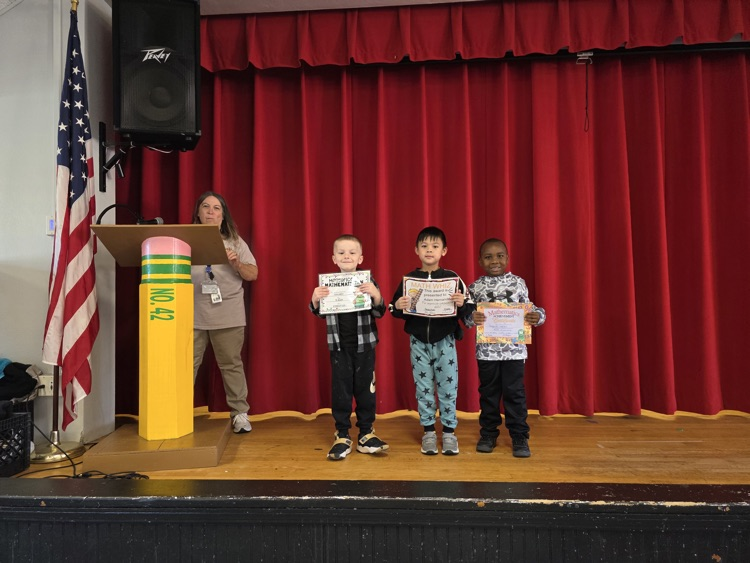 Four children on stage holding certificates, a woman at a podium, American flag and speaker on stage.