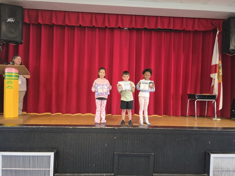 Four children on stage hold certificates, standing on a wooden floor with red curtains behind them.