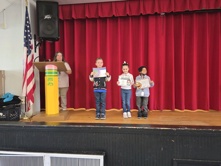 Four children stand on a stage holding certificates, with a woman at a podium and an American flag.