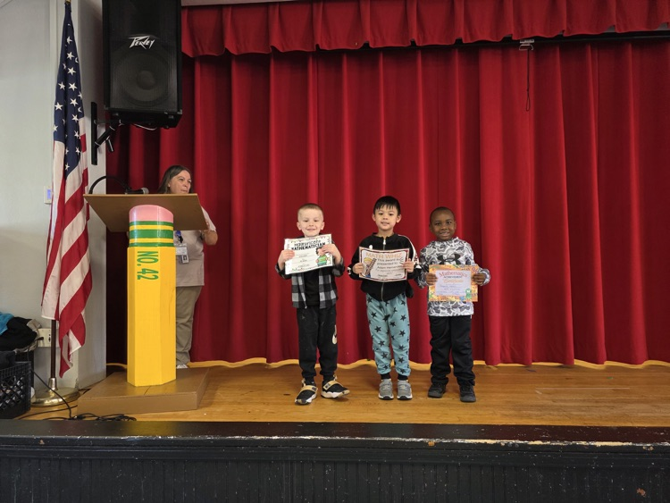 Four children stand on stage holding certificates. A podium with a microphone is behind them. An American flag and trash can are on the left.
