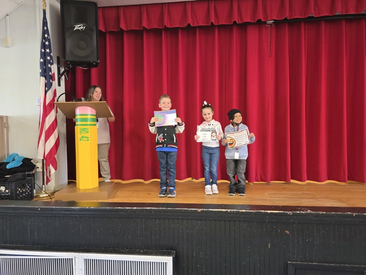 Four children holding certificates stand on stage in front of a woman and an American flag.