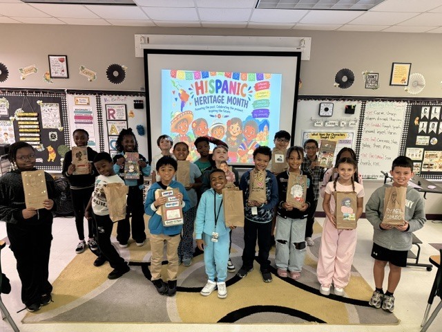 Group of students holding paper bags standing in a classroom, with a projector screen showing Hispanic Heritage Month.