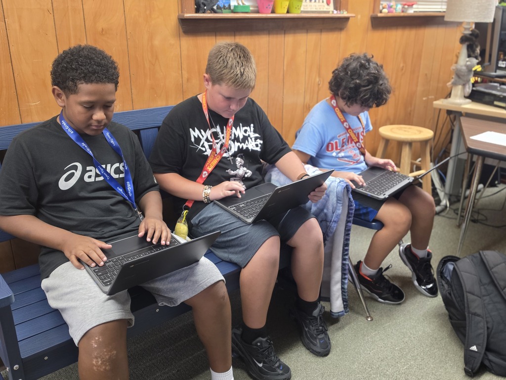 Three students sit on a bench, each using a laptop. Two have lanyards. A backpack rests on the floor behind them.