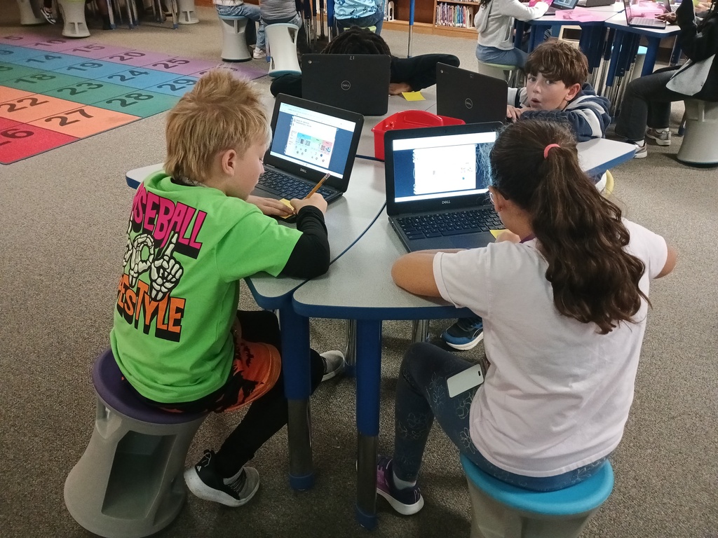 Three students seated on stools at a desk use laptop computers. Rug with letters and numbers on floor.