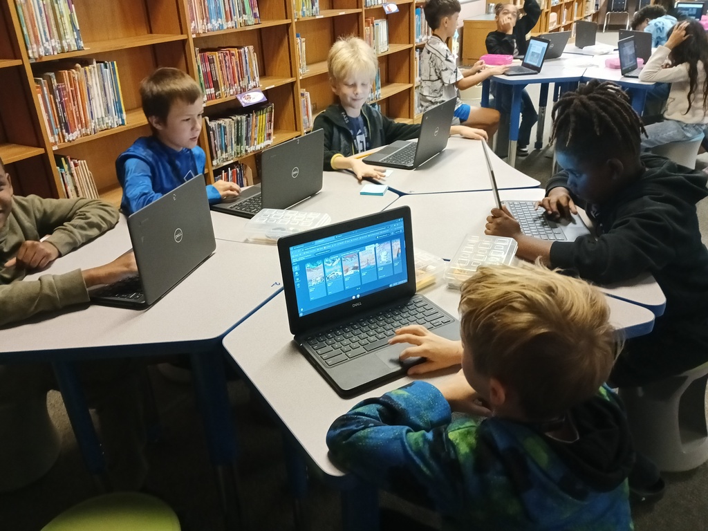 Several students use laptops in a classroom; shelves filled with books in the background.