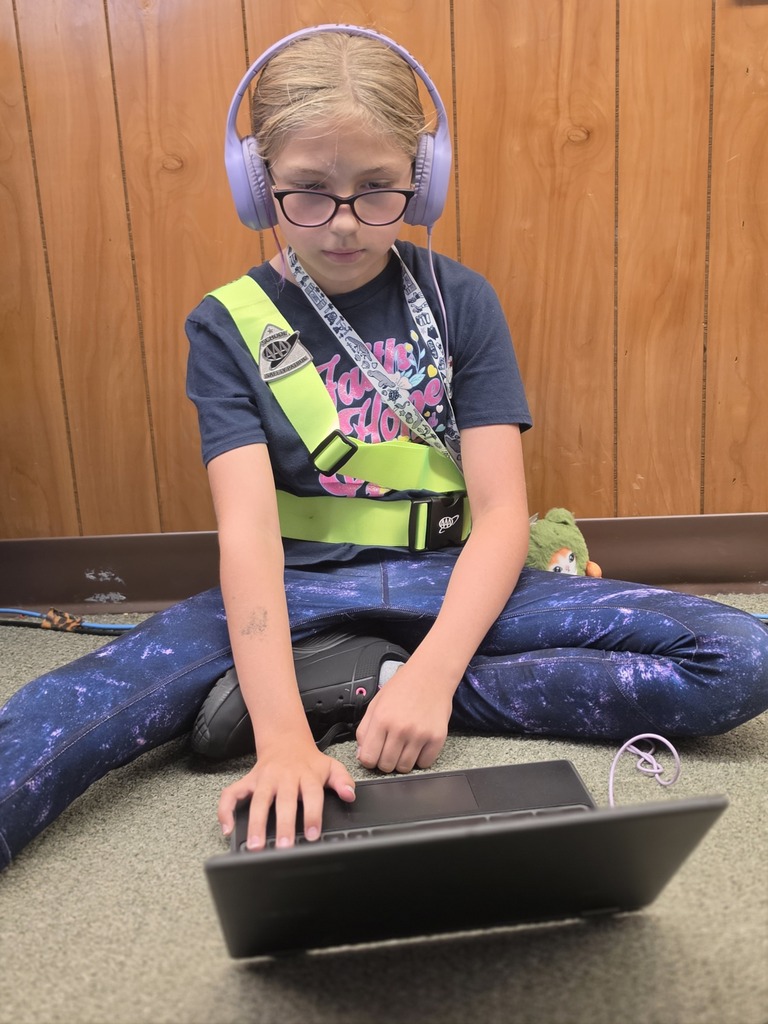 Child wearing glasses, headphones, and a safety vest seated on floor using a laptop.