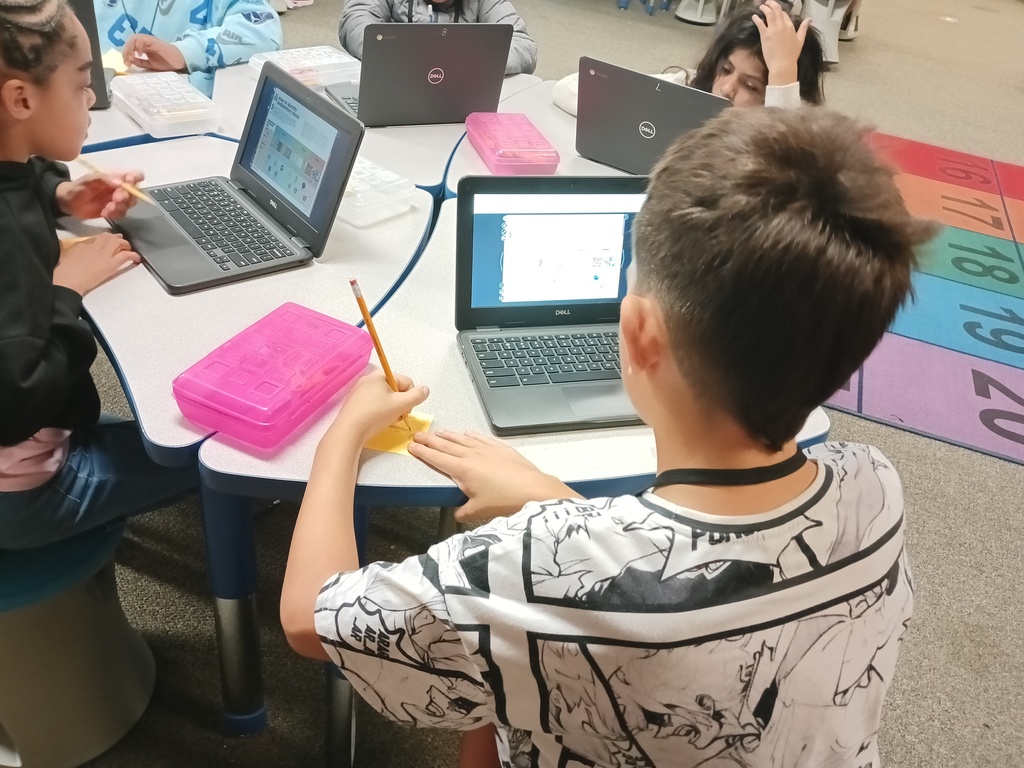 Children in classroom seated at tables with laptops; one holds pencil and pen, another holds pencil.