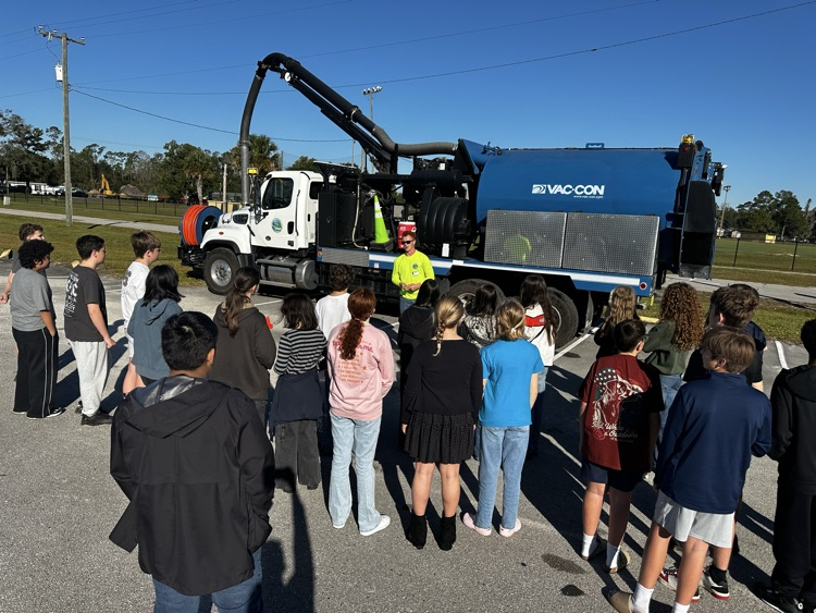 A group of students in front of a garbage truck, with a person in a yellow jacket.