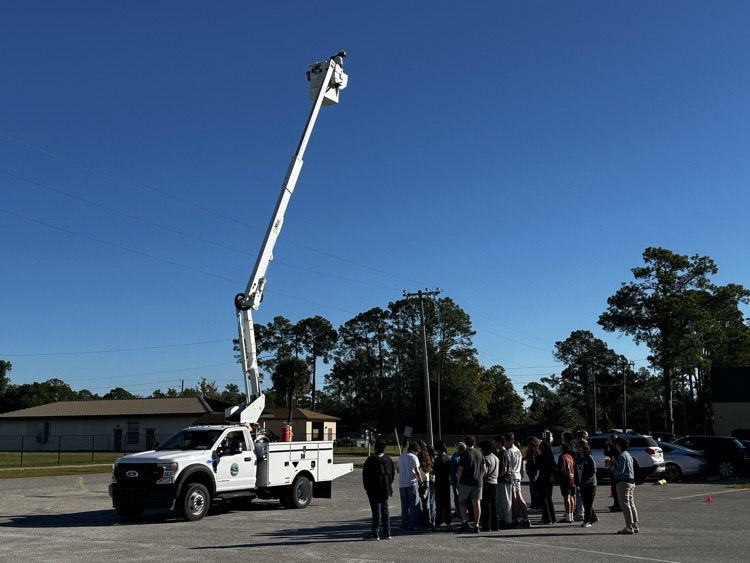 A white truck with a raised bucket is in a parking lot. People stand in a group.