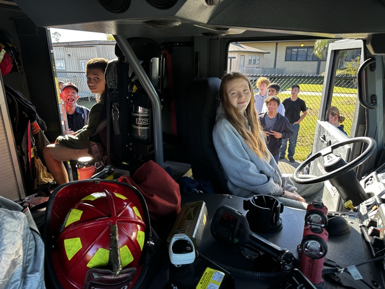 Students sit in fire truck; one in driver seat, others look at items. Helmet, equipment, and building in background.
