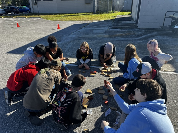 Students sitting in a circle on a parking lot, engaging in an activity with small blocks.